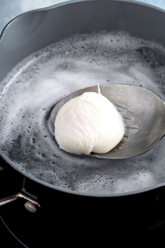 A slotted spoon lifting a poached egg from a pot with hot poaching water.