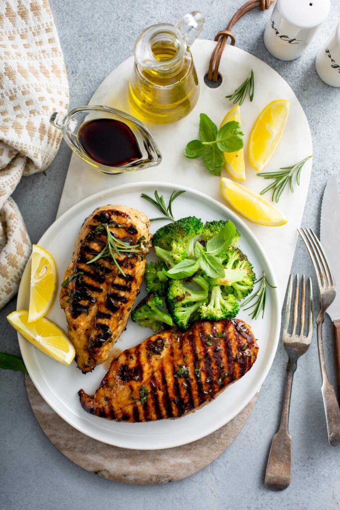 Top view of 2 Grilled Chicken with Balsamic Marinade on a white plate and garnished with slices of lemon and chopped broccoli, next to an olive oil and a balsamic cruet