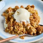 Homemade apple crisp with golden brown oat topping and scoops of vanilla ice cream in a baking dish.