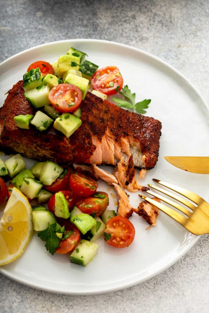 Flaky salmon fillet with blackening spices on a white plate topped with cucumber, tomatoes and avocado salad