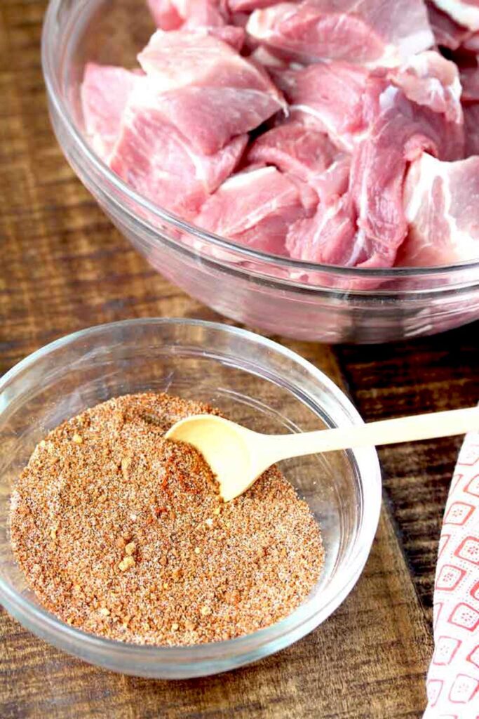 Small glass bowl filled with homemade spice mix next to a bowl of pork cut into big chunks