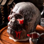 Chocolate cake shaped like a skull and coated with vanilla glaze on a wooden plate.