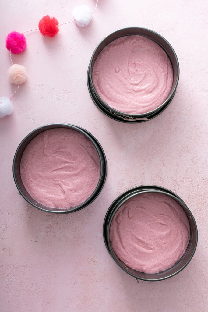 Three 8-inch round baking pans filled halfway with pink cake batter.