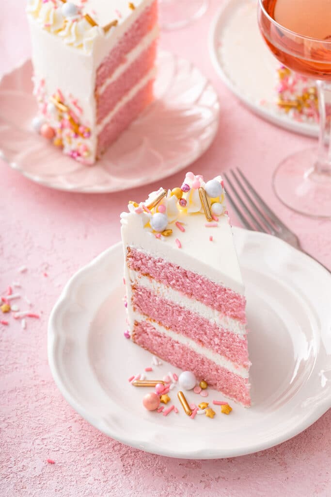Slices of three layer pink champagne cake frosted with Swiss buttercream and decorated with white, gold and pink sprinkles served on white plates.
