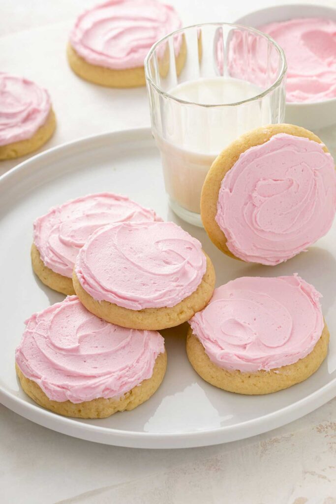 Pink frosted soft sugar cookies on a plate next to a glass of milk.