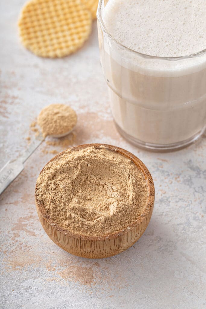 A small wooden bowl filled with maca root powder next to a glass of maca latte.