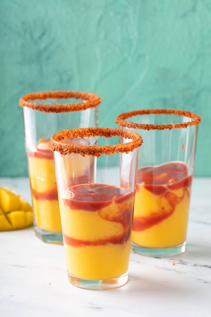 three tall glasses with tajin on the rim and half full with mango smoothie and chamoy sauce