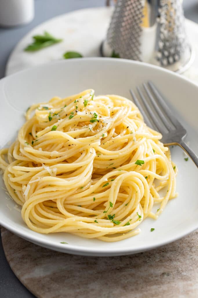 A dinner plate with a serving mound of silky garlic butter pasta sprinkled with freshly grated Parmesan cheese and garnished with chopped fresh parsley.