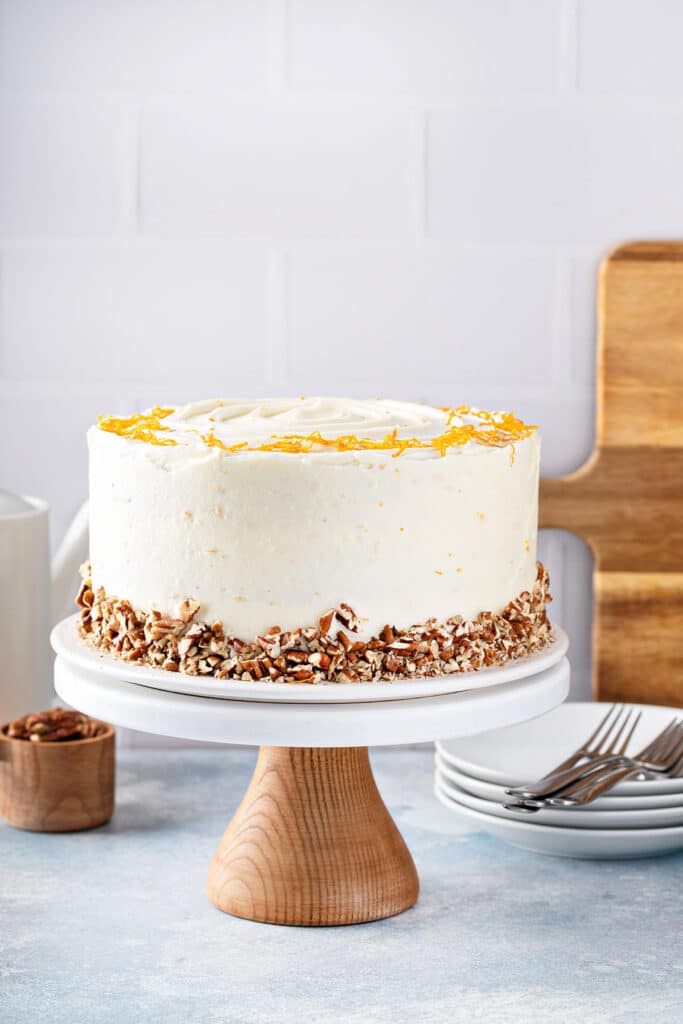 Cream cheese frosted carrot cake decorated with chopped pecans and orange zest on a cake stand.