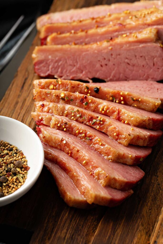 Slices of meaty and tender corned beef next to a small bowl of corned beef pickling spices on a wooden board.
