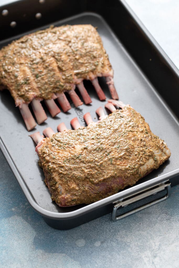 Two racks of lamb coated with an herb and Dijon mustard mixture in a roasting pan.