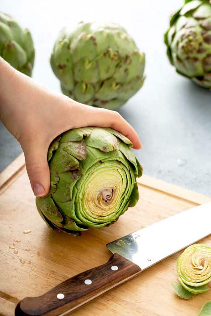 holding an artichoke with the stem cut off on a cutting board