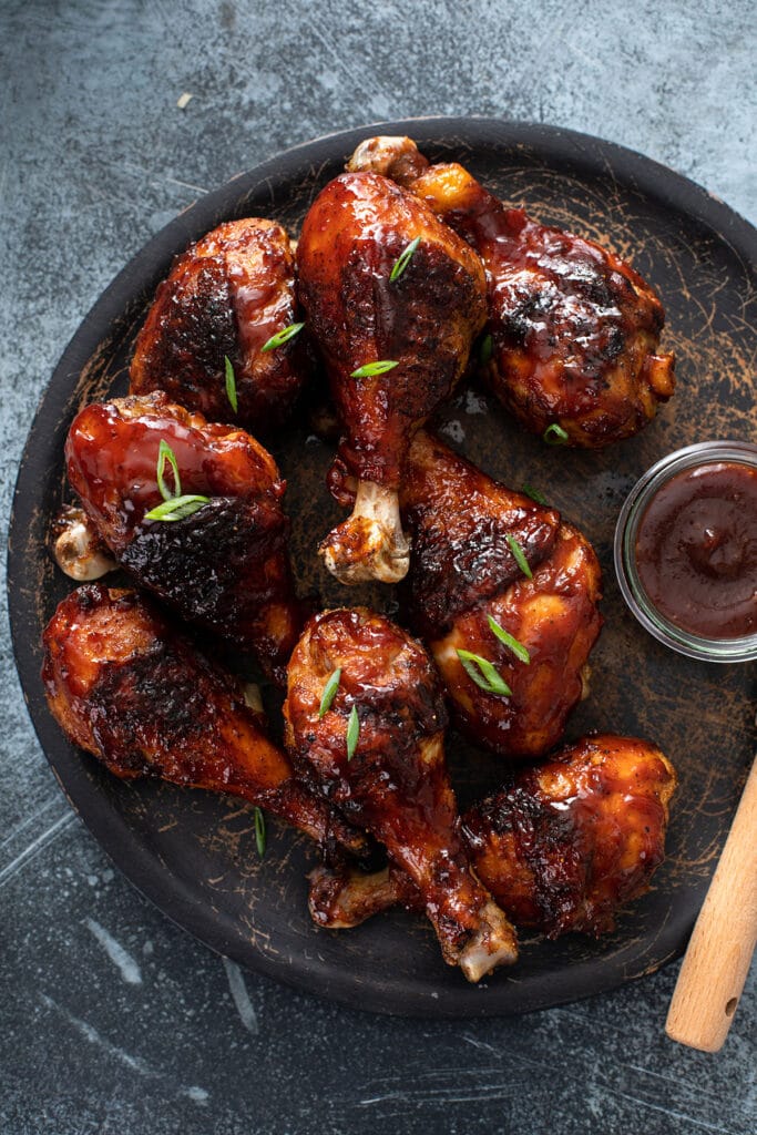 Chicken legs grilled to golden perfection and smothered in BBQ sauce on a black plate.