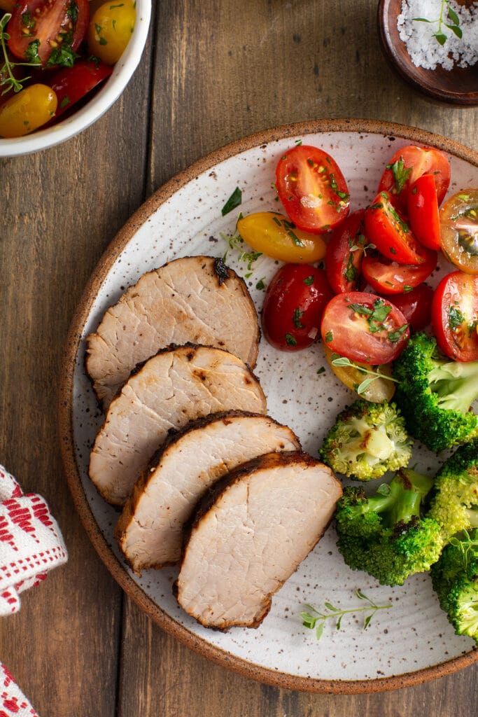Sliced pork tenderloin served with fresh tomatoes and steamed broccoli in a white plate.