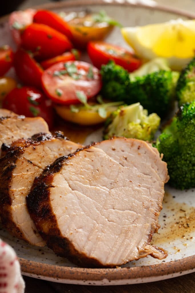 Close up view of a few slices of grilled pork tenderloin on a plate with broccoli and cherry tomatoes