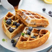 Maple syrup pouring over lemon blueberry waffles topped with fresh blueberries, sliced almonds and lemon zest.