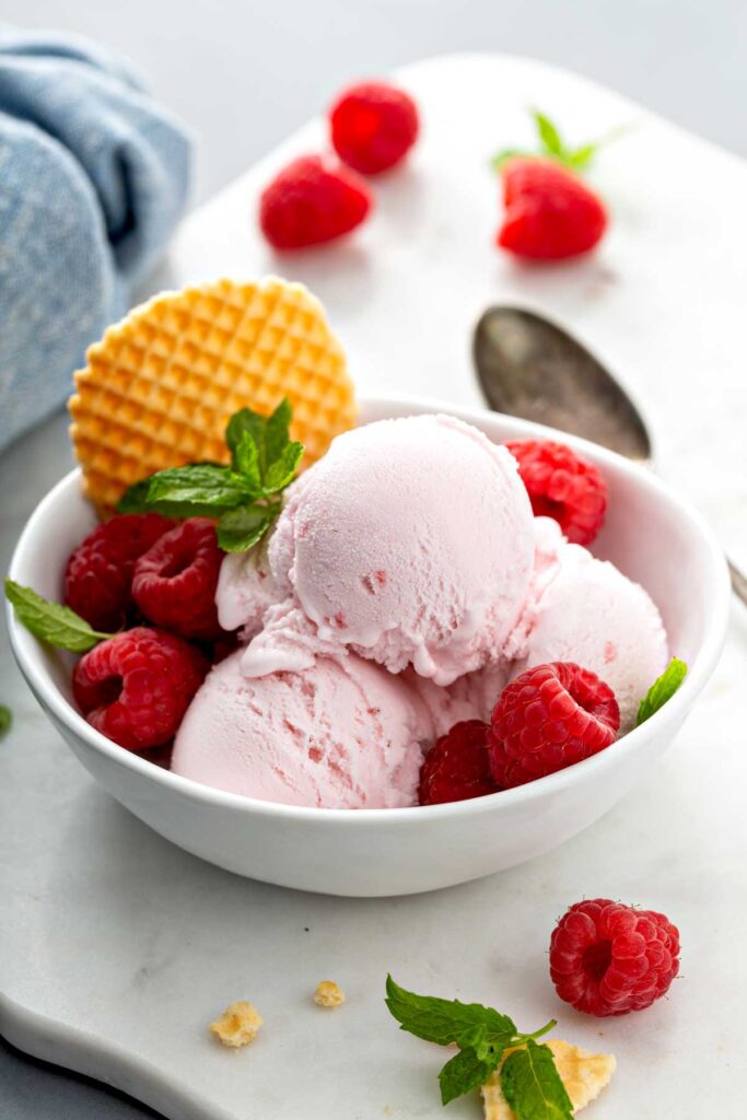 Three scoops of raspberry ice cream in a white bowl garnished with fresh raspberries and a waffle cookie.