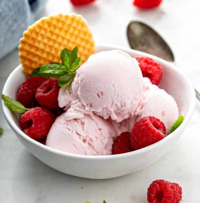 Three scoops of raspberry ice cream in a white bowl garnished with fresh raspberries and a waffle cookie.