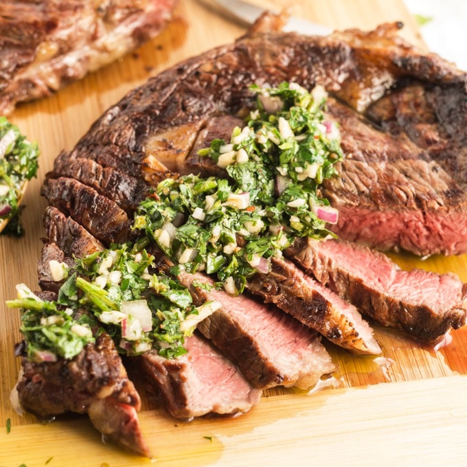 sliced grilled ribeye steak topped with chimichurri sauce on a wooden cutting board
