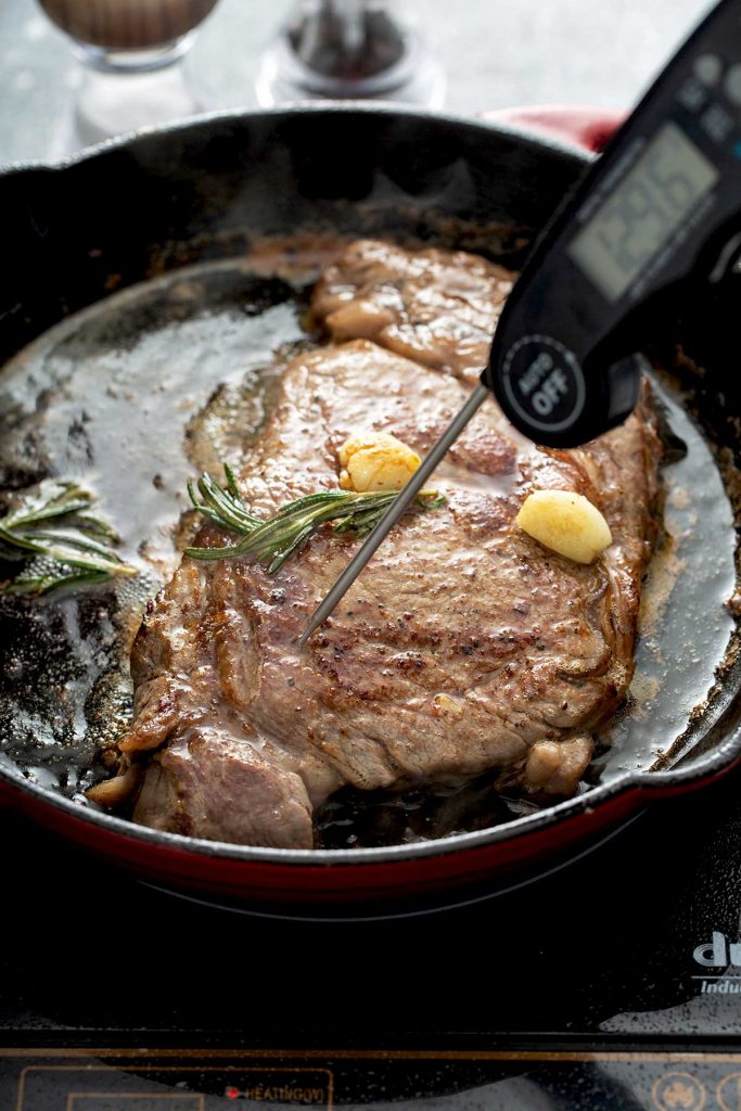 Checking the internal temperature of a juicy ribeye steak cooking in a skillet with garlic and rosemary.