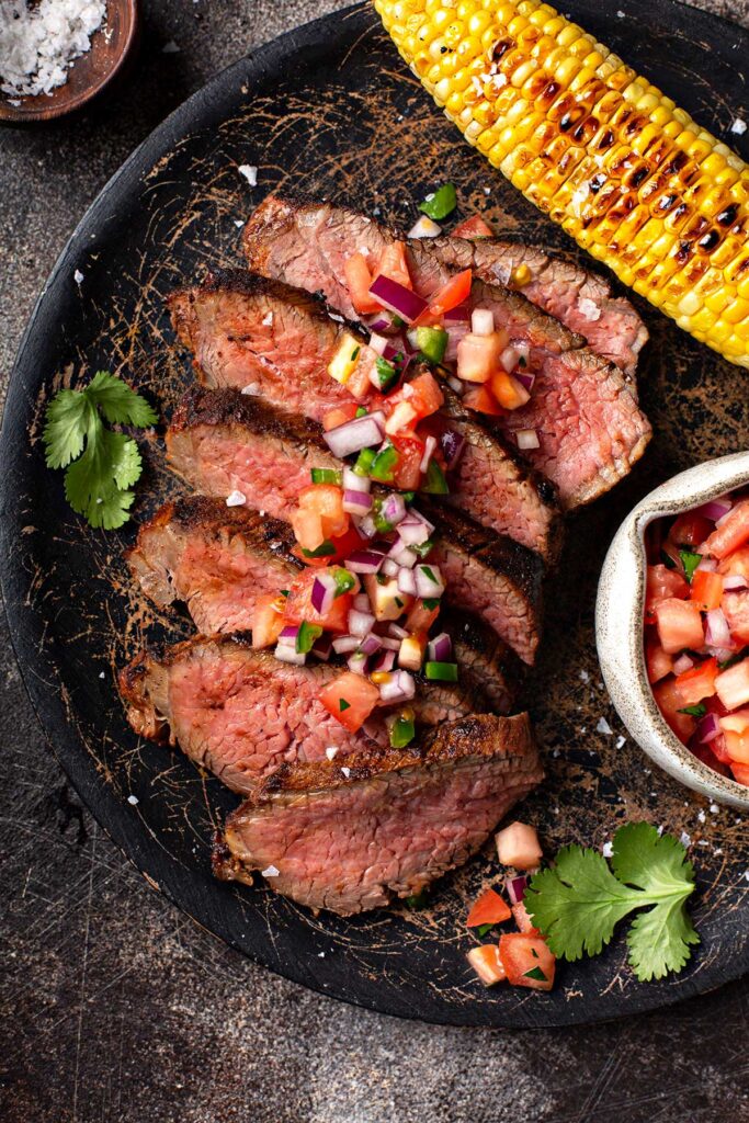 A plate with slices of tri tip topped with pico de gallo and served with grilled corn on the cob.