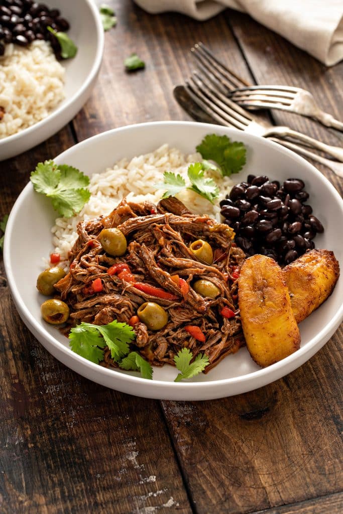 A plate filled with ropa vieja beef, black beans, white rice and fried maduros (friedripe sweet plantains).