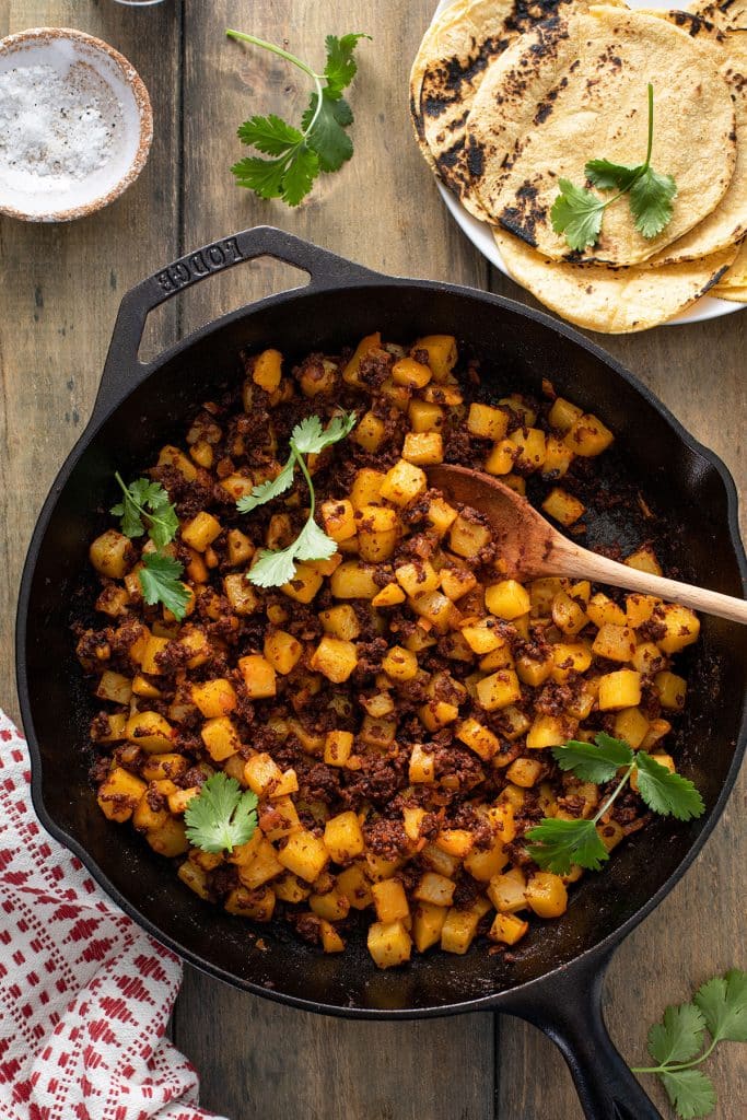A cast iron skillet with a wooden spoon serving potatoes and Mexican chorizo