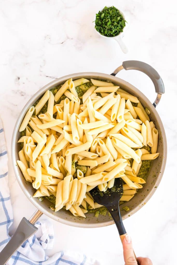 mixing cooked pasta with the pesto sauce in the skillet