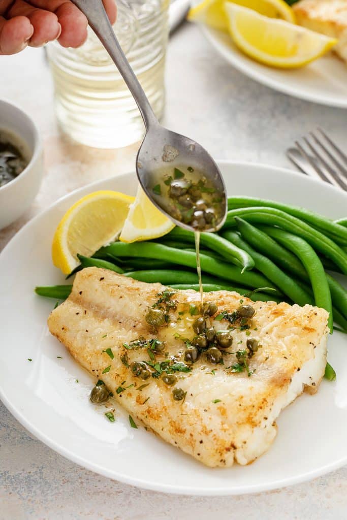 Pouring lemon butter with capers over lightly golden pan seared cod fillet.