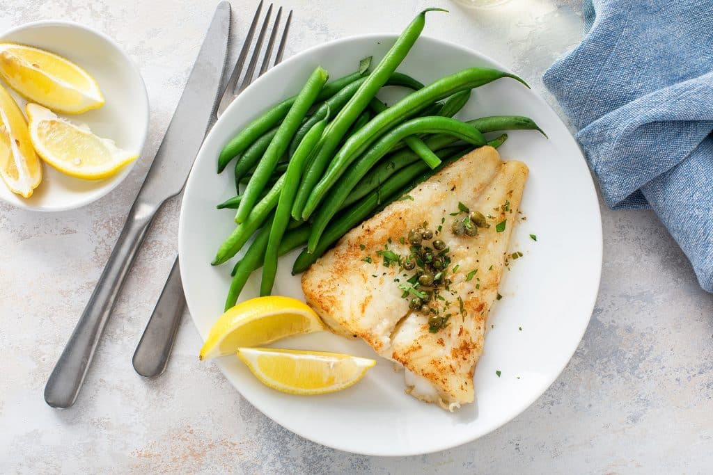 Pan seared cod smothered in lemon butter served with lemon wedges and green beans on a white plate.