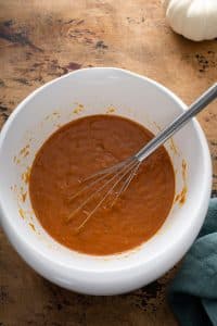 oil, pumpkin puree, brown sugar, granulated sugar, eggs, and vanilla extract in a white bowl with a whisk