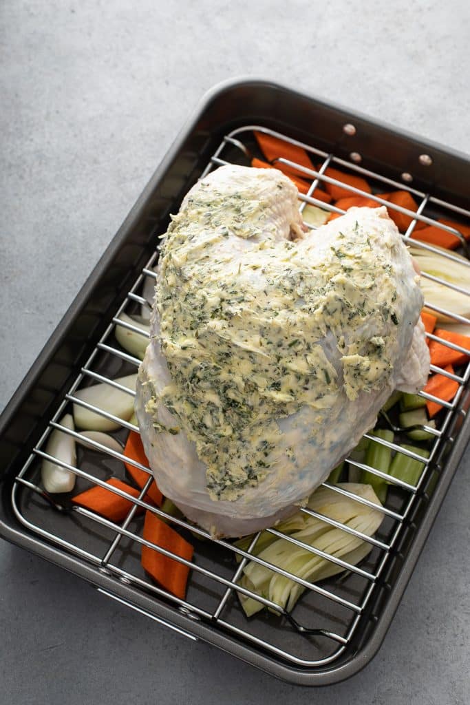 Turkey breast slathered with herb butter on a rack over vegetables in a roasting pan