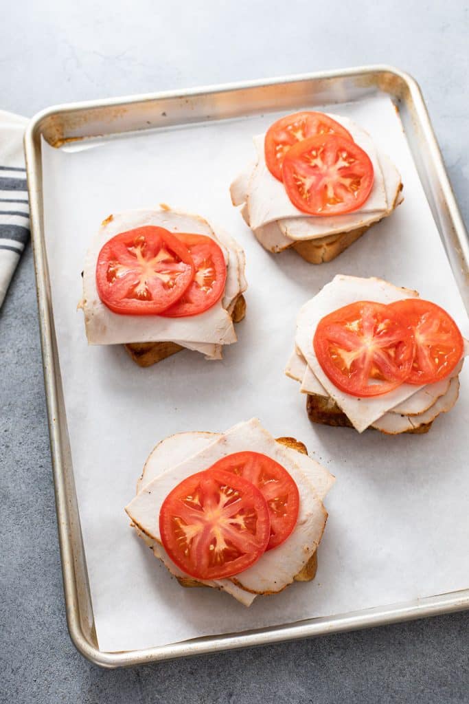 Toasted bread topped with sliced turkey and sliced tomato on a baking sheet pan