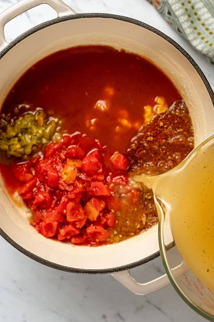 pouring broth over green chiles, tomatoes, spices, seasonings and enchilada sauce