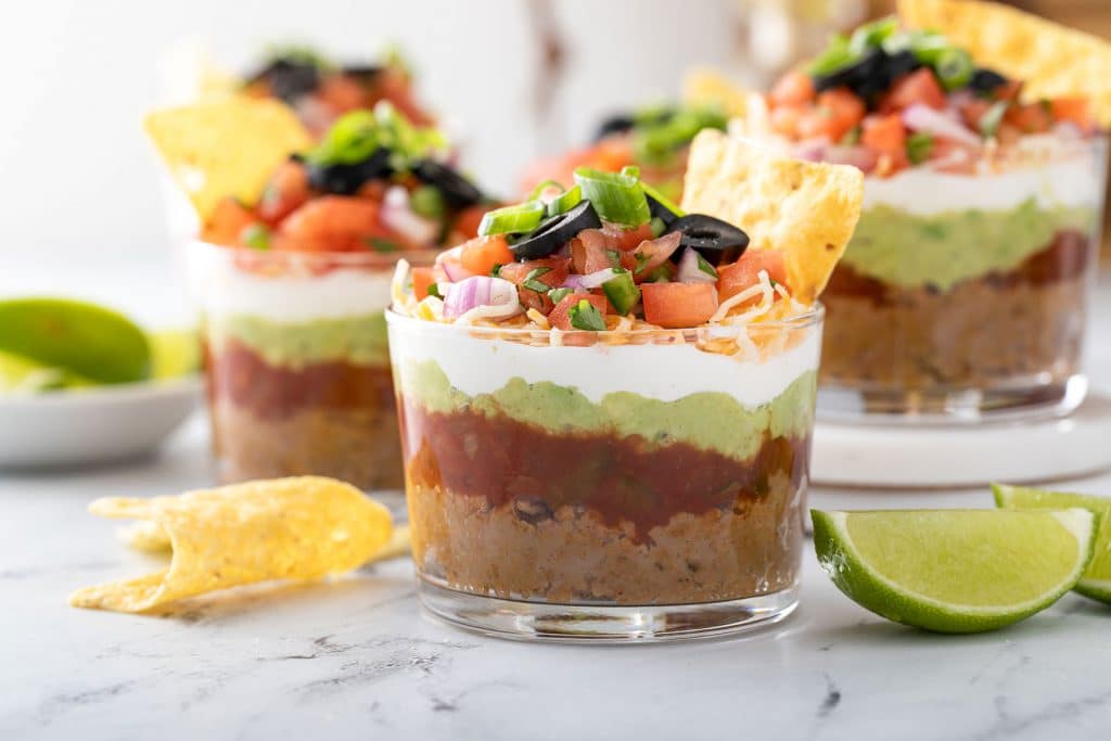 Clear cups with layers of refried beans, salsa, guacamole, sour cream, shredded cheese, pico de gallo, olives and green onions.