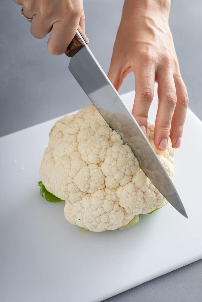 A whole cauliflower on a cutting board getting cut in halsf