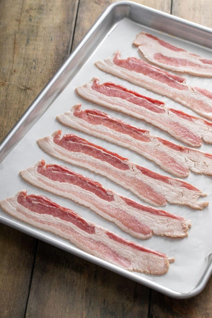 Raw bacon strips on a parchment paper lined baking sheet.