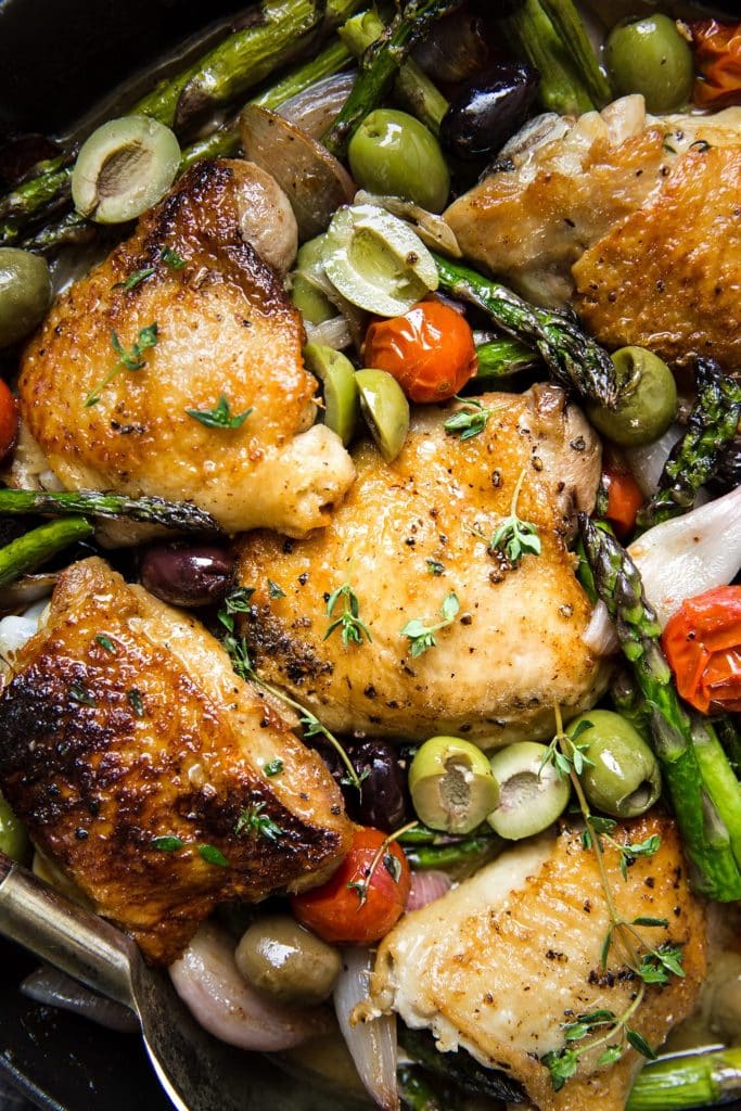 Golden brown chicken thighs in a skillet with olives, tomatoes and asparagus