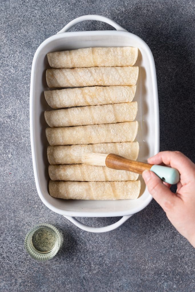 Rolled up corn tortillas in a baking dish getting brushed with oil