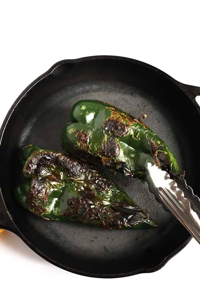 two roasted poblano peppers in a cast iron skillet being flipped with tongs 
