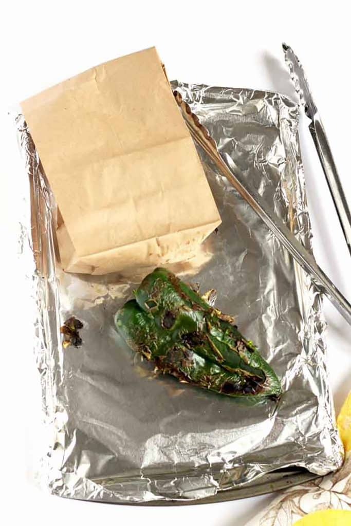 roasted poblano pepper on a sheet pan next to a paper bag and tongs