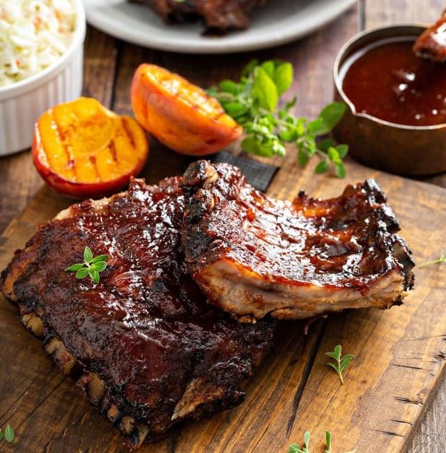 baked ribs cut up into 3 ribs each on a wooden board, with bbq sauce and grilled peaches.