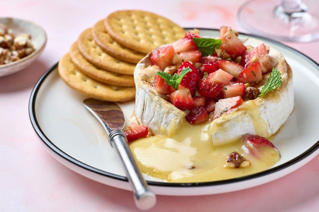 A round of baked brie cheese with nuts topped with strawberries and surrounded by crackers on a white plate.