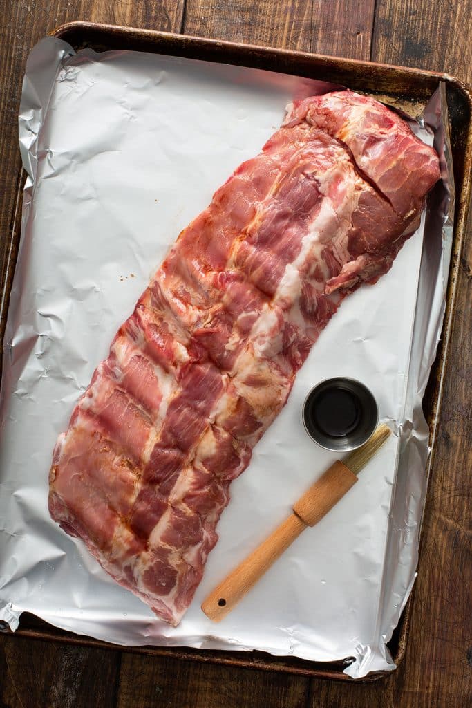 A rack of ribs, a brush and a small bowl with liquid smoke on a sheet pan.
