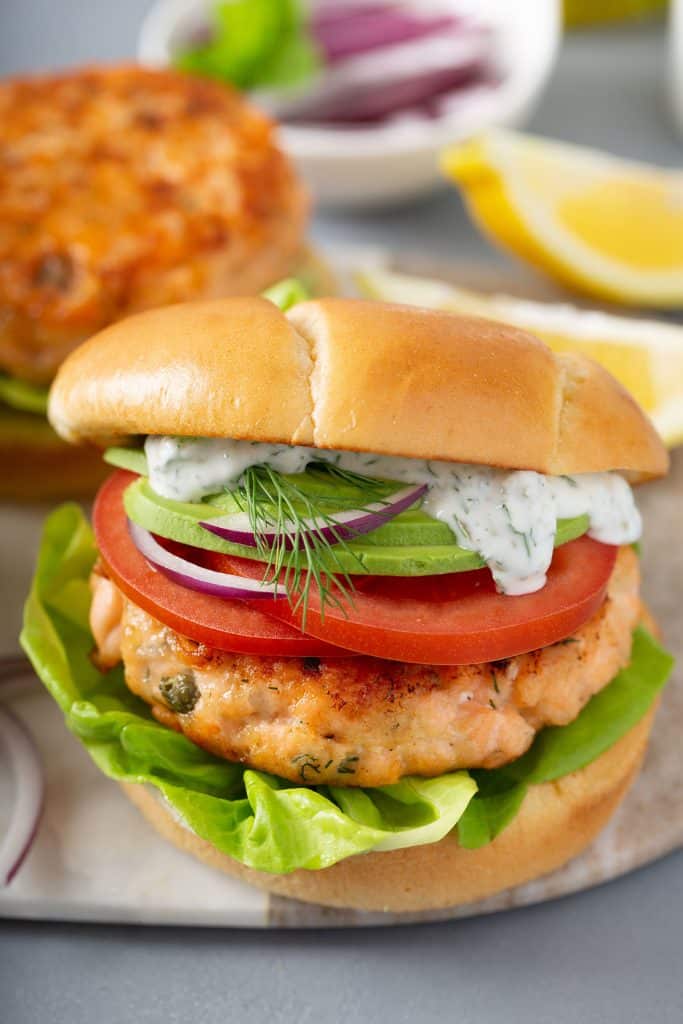 Salmon burger on a bun with creamy sauce, sliced tomatoes, red onion slices and avocado slices.