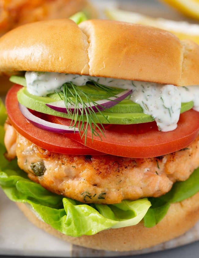Salmon burger topped with tomato slices, sliced red onions, sliced avocado and a creamy sauce inside a brioche burger bun