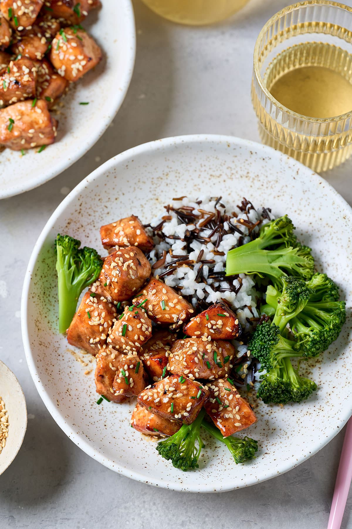 Glazed salmon bites topped with sesame seeds and chives, served over a wild rice blend with steamed broccoli in a speckled bowl. A second plate of salmon and a glass of white wine sit alongside.