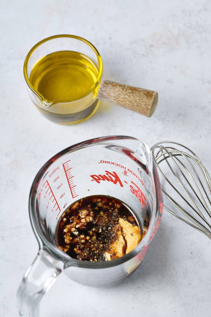 glass of oil and large measuring cup of ingredients next to whisk