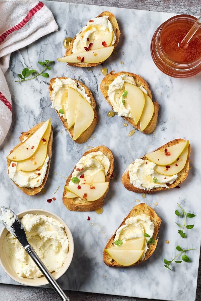 seven mini toasts with ricotta cheese, slices of pear and drizzled with hot honey, over a marble surface
