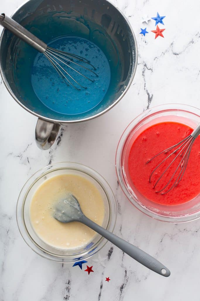 Cake batter divided into 3 bowls. One is colored blue, the other red and the third one left as it is.
